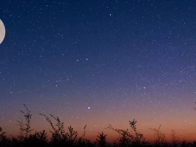 Full moon over a field amidst a purple and orange starry night sky